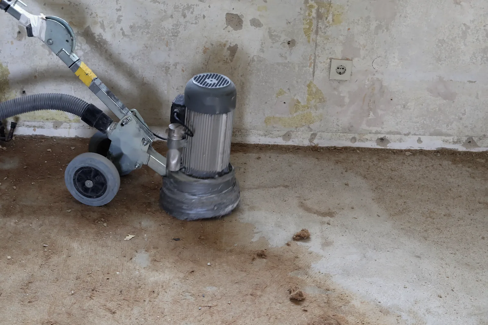Schleifmaschine mit Absaugschlauch auf dem Estrich vor einer alten Wand mit Steckdose