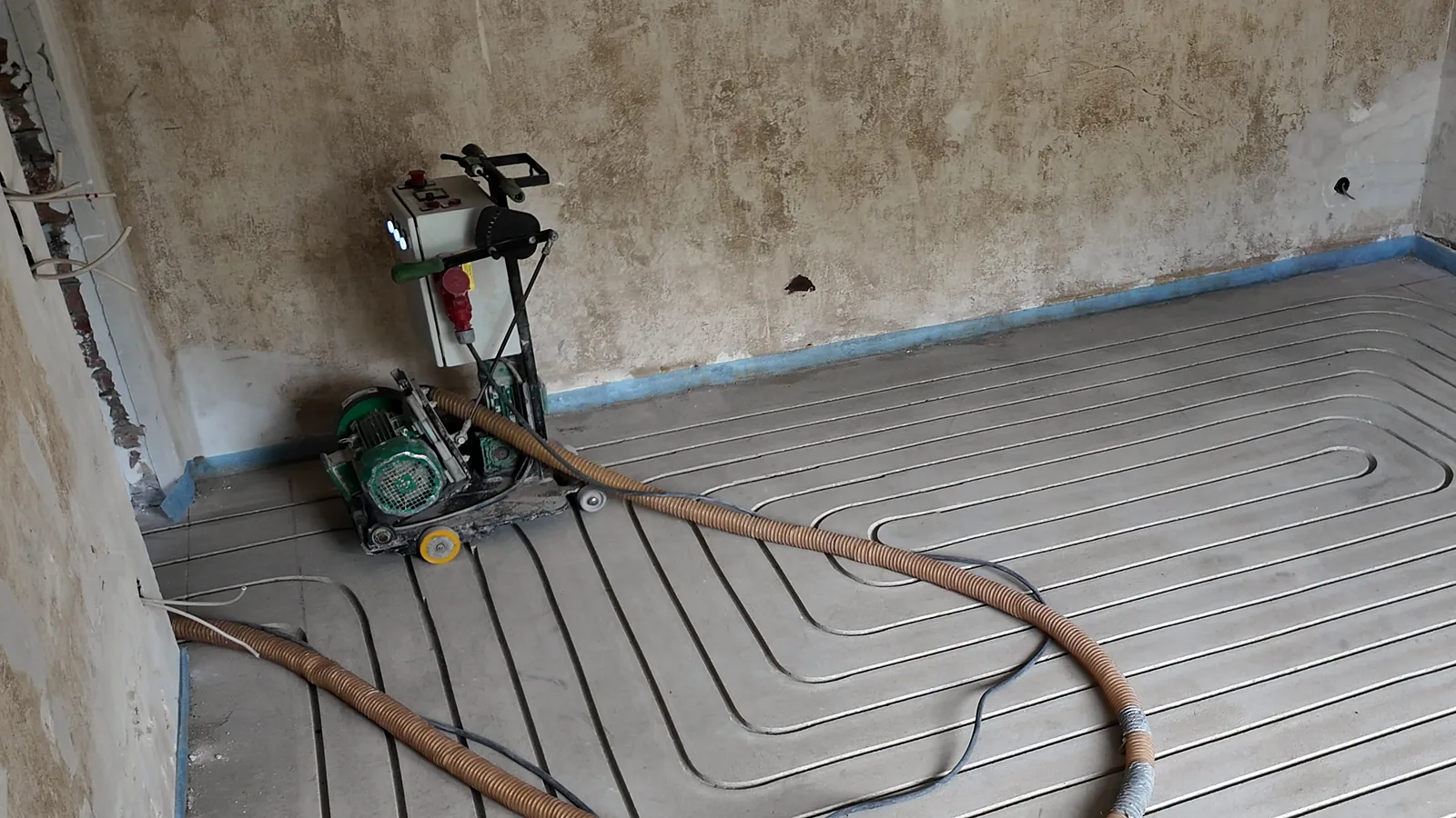 Fräsmaschine mit Schaltkasten an der Wand, im Boden daneben Kanäle mit Heizrohr, ein Absaugschlauch liegt quer