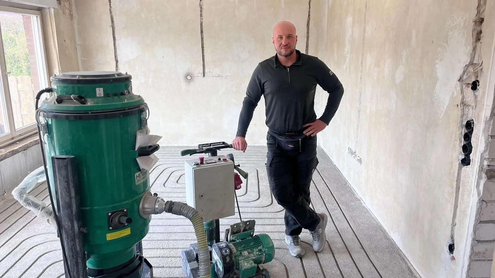 Kamil Bregulla im entkernten Raum neben der Fräsmaschine, im Estrich die gefrästen Kanäle für die Fußbodenheizung